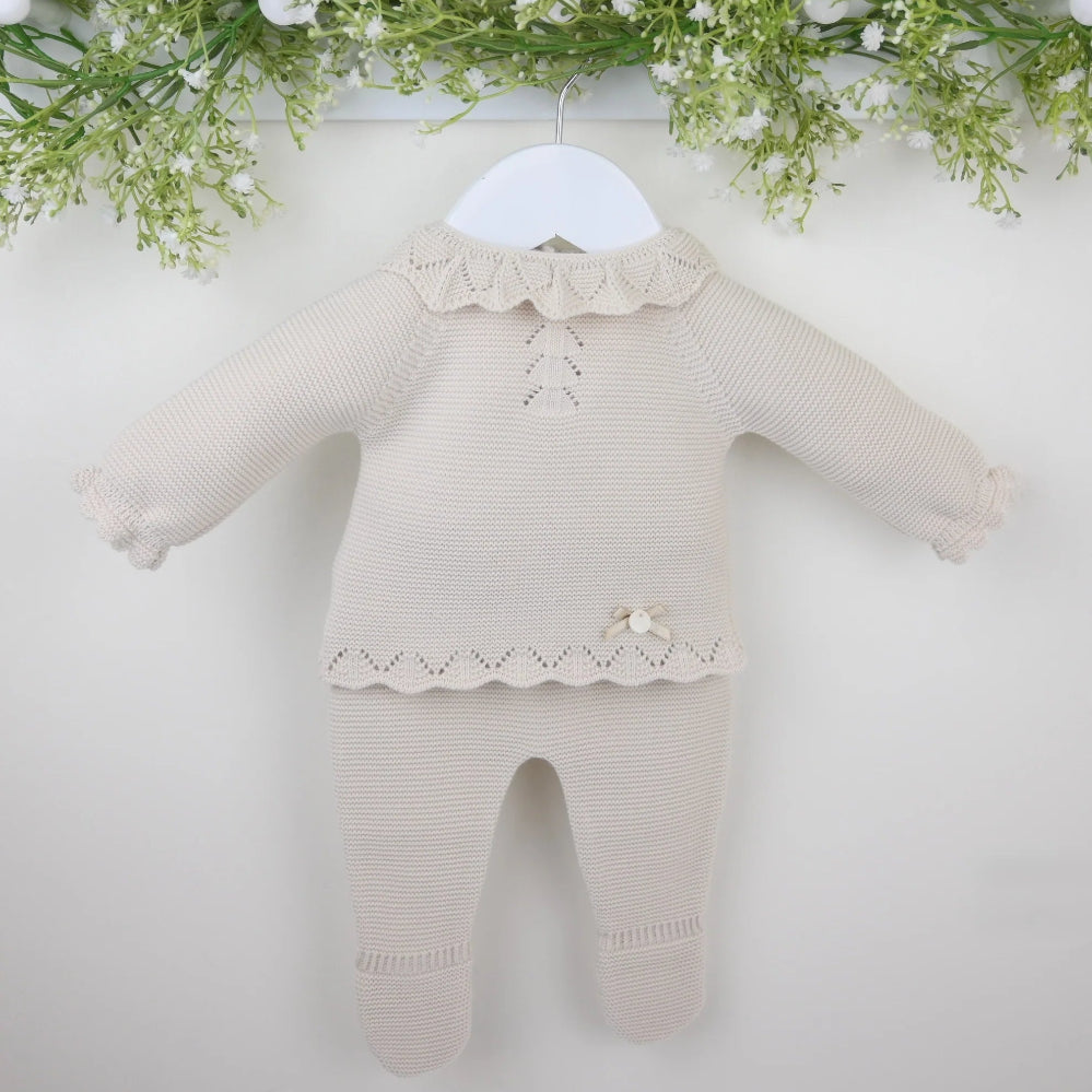 A beige knitted baby outfit consisting of a top with a textured frilled collar and chevron patterned chest, matching trousers with enclosed feet, and a bonnet, displayed on a white hanger against a floral backdrop.