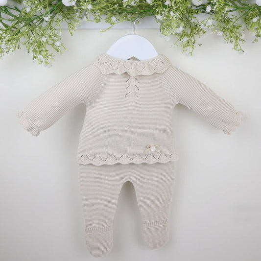 A beige knitted baby outfit consisting of a top with a textured frilled collar and chevron patterned chest, matching trousers with enclosed feet, and a bonnet, displayed on a white hanger against a floral backdrop.